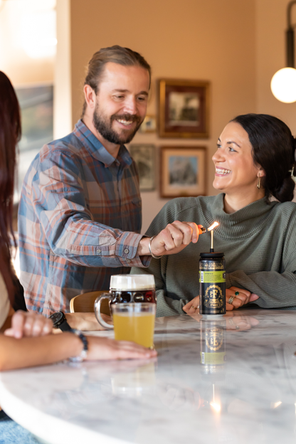 Two people sitting at a table with drinks, one lighting a candle.