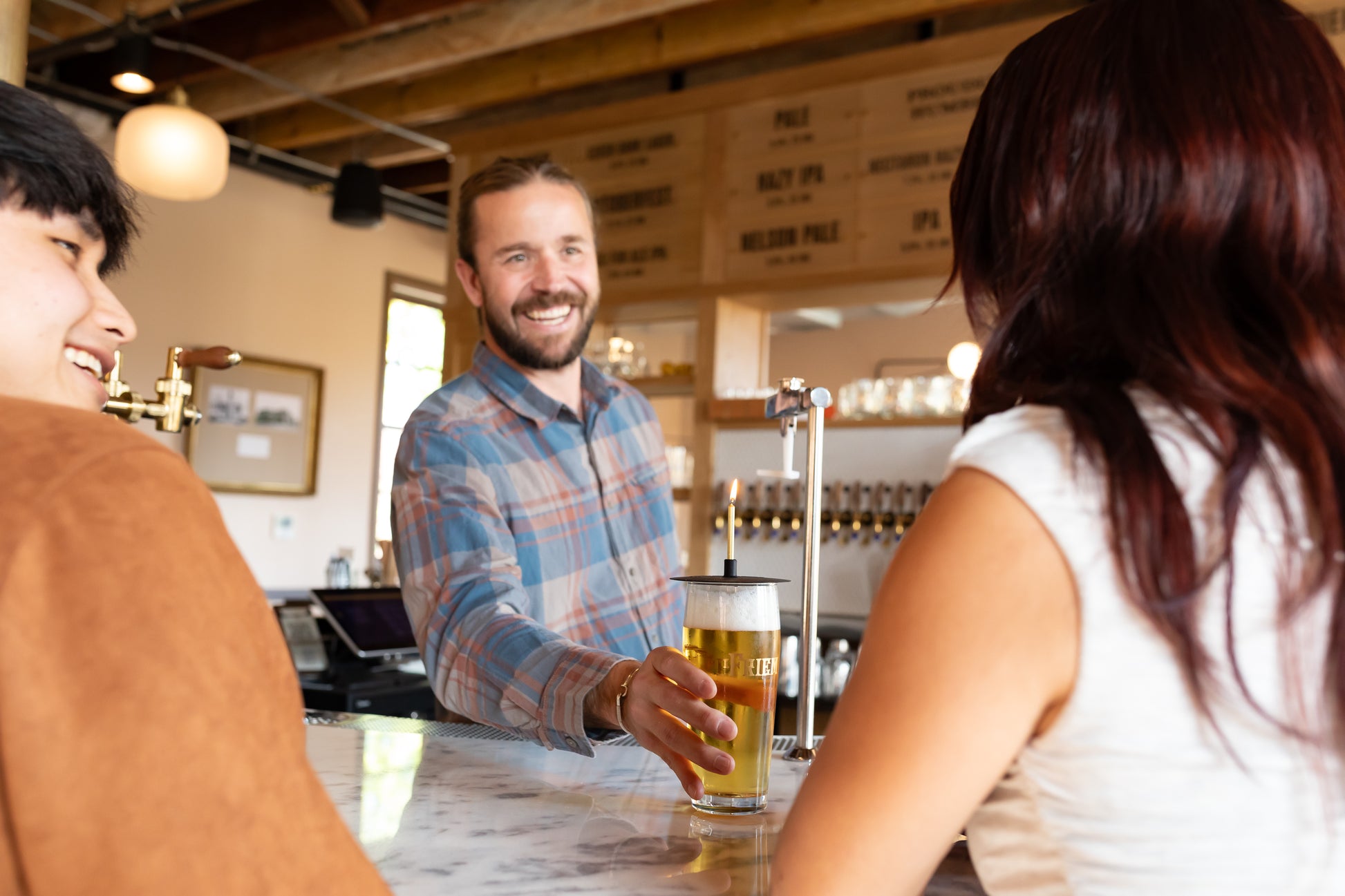 Brewery Bartender serving a birthday beer drink to two customers in a cozy bar setting.