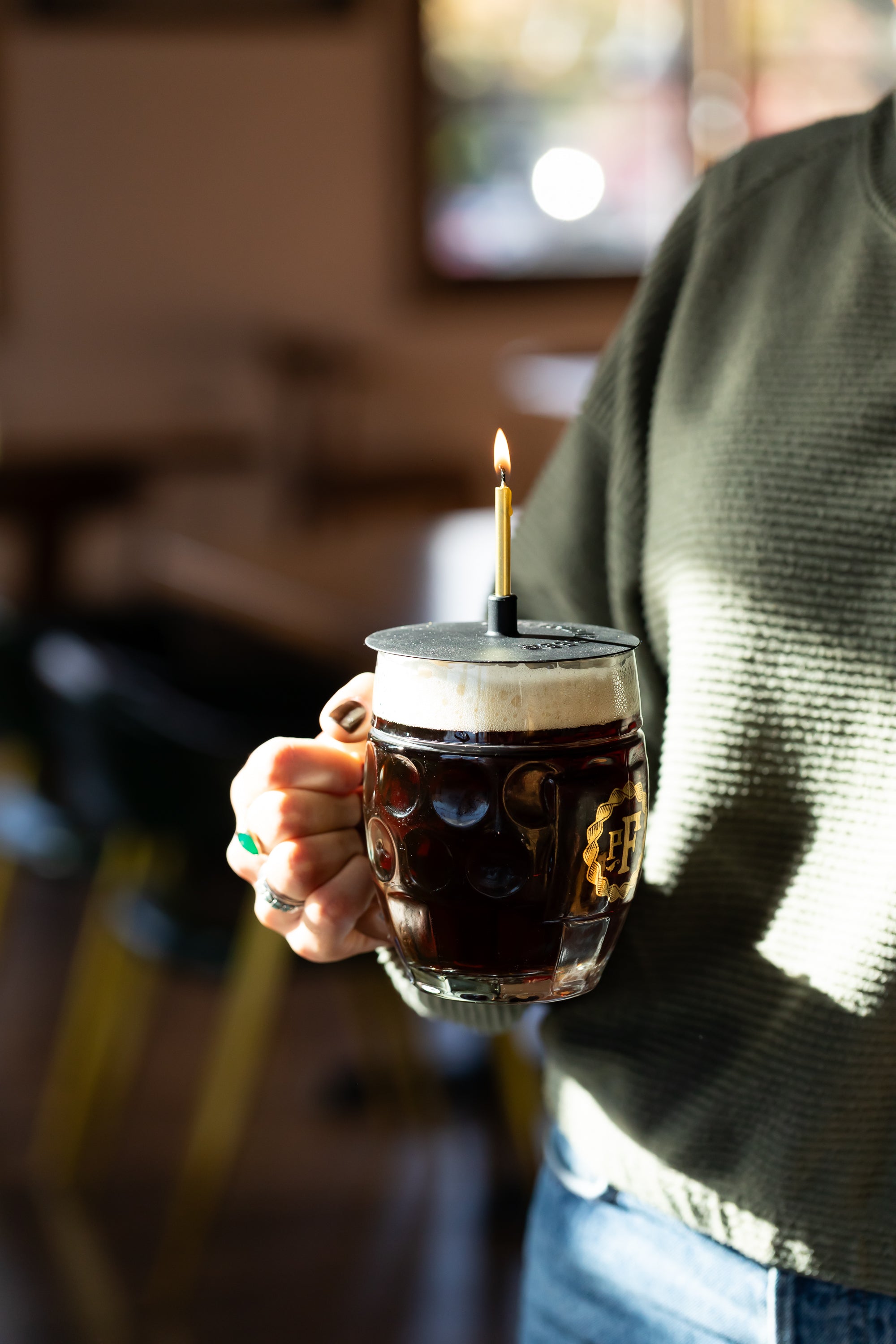 Person holding a mug of dark beer with a lit candle on top, blurred background